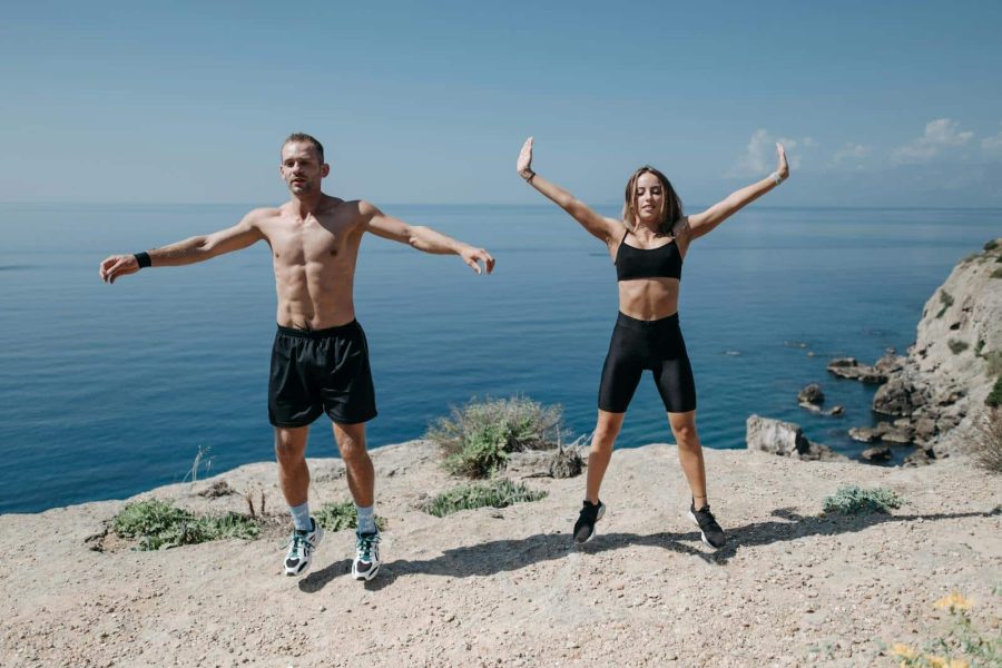 A man and woman perform jumping jacks on a cliff by the ocean under a clear sky.