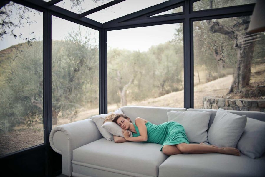 A woman in green dress naps on a sofa in a glass-walled room with olive trees outside.